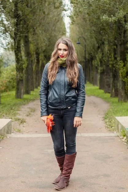 A woman wearing a black quilted vest over a white button-down shirt. She is also wearing dark-wash jeans and brown ankle boots. The background is a blurred outdoor setting.