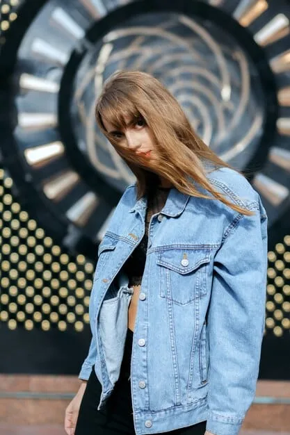 Gigi Hadid is photographed in a medium shot, wearing a light wash denim jacket over a plain white t-shirt, paired with slightly darker wash jeans. She accessorizes with white sneakers and sunglasses. Her hair is styled in loose waves. The background is a blurred city street.