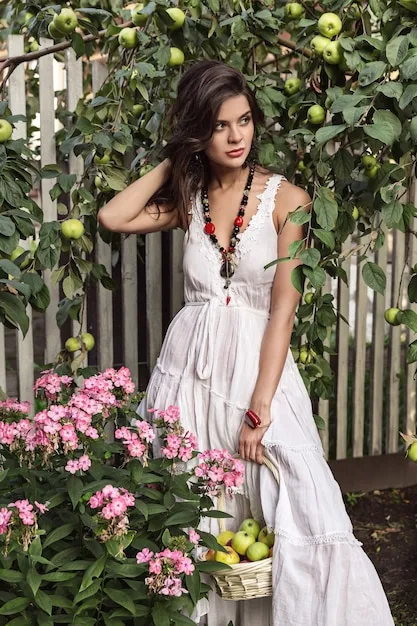 A woman with long, brunette hair, styled in loose, flowing waves. She is wearing a boho-chic dress and standing in a sunny garden.