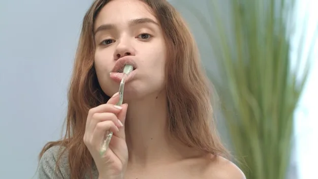 A woman gently exfoliating her lips with a soft toothbrush, followed by the application of a clear lip balm. The focus is on the gentle circular motions of the toothbrush and the smooth texture of the balm.