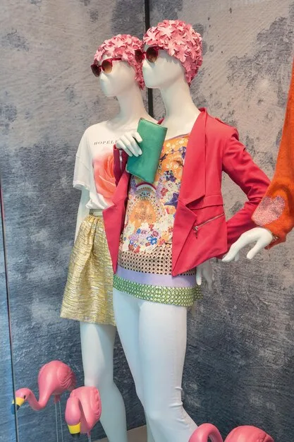 A mannequin in a department store window dressed in a vibrant color-blocked outfit - a pink blazer, green pants, and a blue top. The window display features other colorful garments and accessories.