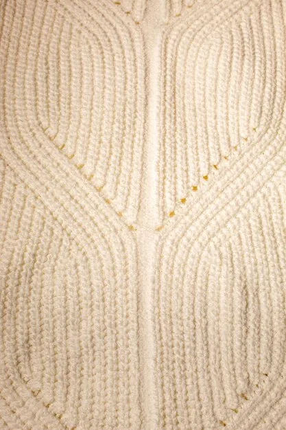 A close-up shot of a hand-knitted wool sweater in a cream color, featuring intricate cable knit patterns. The background is blurred, showing a cozy, rustic interior with a wooden table and soft lighting.
