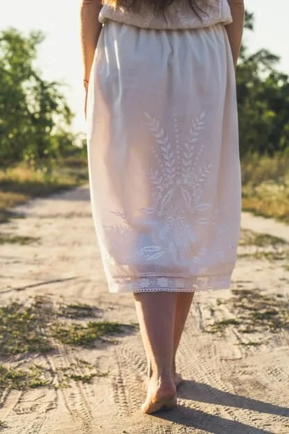 A woman wearing a long, flowy linen dress with a delicate floral embroidery detail, standing in a field of wildflowers. She is holding a straw hat in one hand and has a serene, content expression on her face. The image is bathed in golden sunlight, creating a warm and inviting atmosphere.