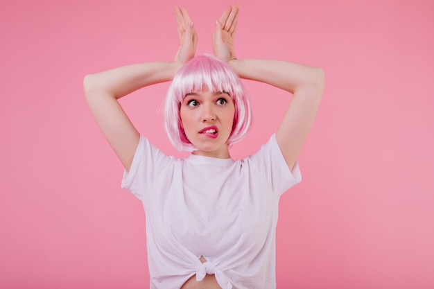 A woman with short, pastel pink hair. The hair is styled in a sleek bob, and she is wearing a white t-shirt. The background is a soft, pastel-colored wall.