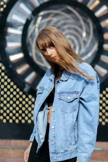Gigi Hadid is photographed in a medium shot, wearing a light wash denim jacket over a plain white t-shirt, paired with slightly darker wash jeans. She accessorizes with white sneakers and sunglasses. Her hair is styled in loose waves. The background is a blurred city street.