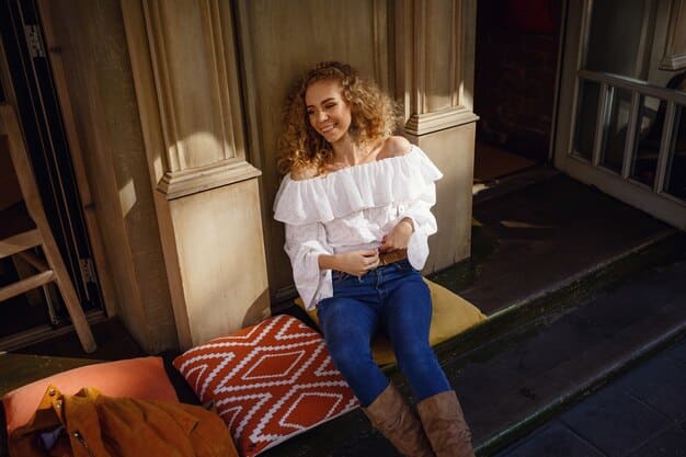 A woman is sitting at an outdoor cafe, wearing dark wash jeans, a cream-colored silk blouse, and ankle boots. She is smiling and looking off to the side, with a coffee cup on the table. The lighting is soft and natural, emphasizing the effortless chic of her outfit.