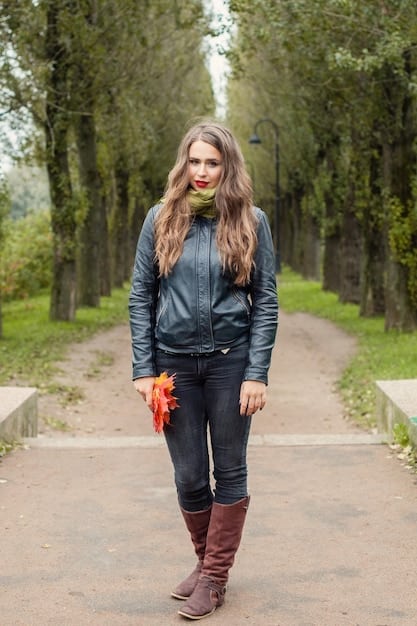 A woman wearing a black quilted vest over a white button-down shirt. She is also wearing dark-wash jeans and brown ankle boots. The background is a blurred outdoor setting.