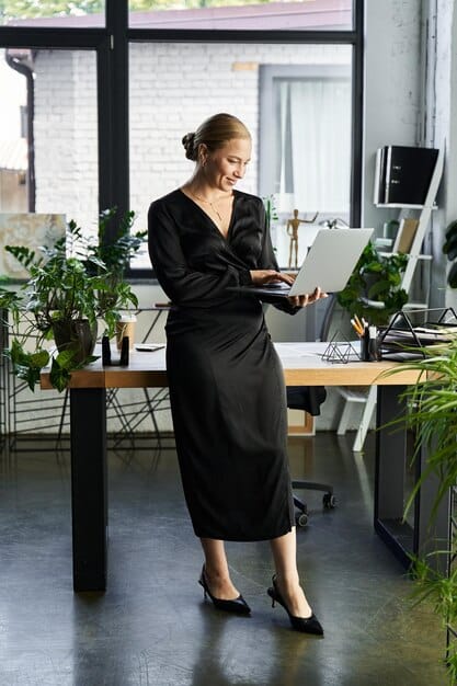 A woman with dark, sleek hair pulled back into a low ponytail. She is wearing a sophisticated black dress and standing in a modern office setting.