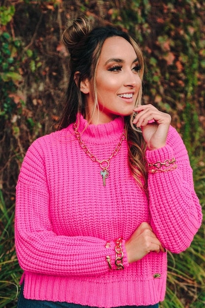 A close-up shot of a woman smiling and wearing a bright yellow oversized sweater with contrasting pink accessories. The background is blurred, emphasizing the outfit.