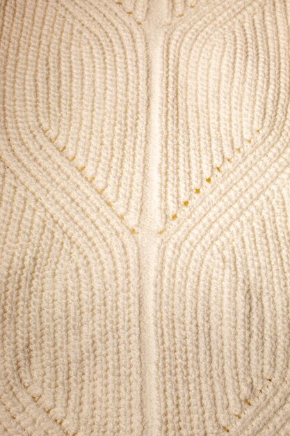 A close-up shot of a hand-knitted wool sweater in a cream color, featuring intricate cable knit patterns. The background is blurred, showing a cozy, rustic interior with a wooden table and soft lighting.