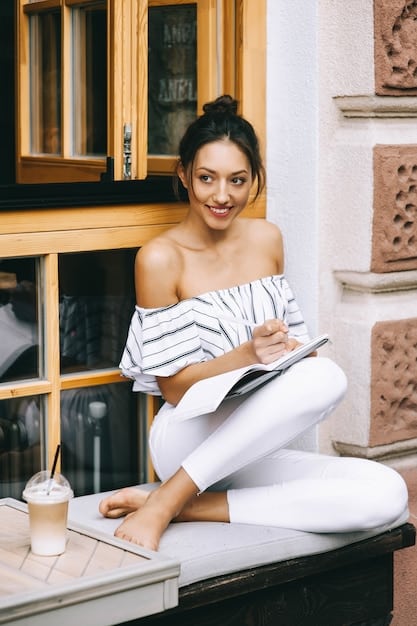 A woman wearing a Shein floral print blouse tucked into black, stretchy trousers, standing in a well-lit home office. She is smiling and holding a pen, creating a professional and approachable image.