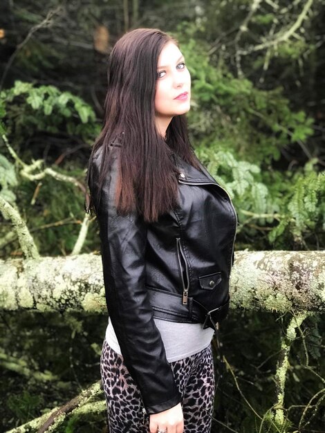 A woman with long brown hair is wearing a black faux leather jacket over a floral midi dress. She also is accessorized with black combat boots and a silver chain necklace. The background is a city park during the early fall with trees showing the first hints of autumn colors.