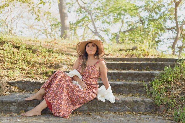 A woman wearing a flowy maxi dress with floral prints from Shein, accessorized with a wide-brimmed hat and layered necklaces, posing in a natural, outdoor setting with warm lighting.