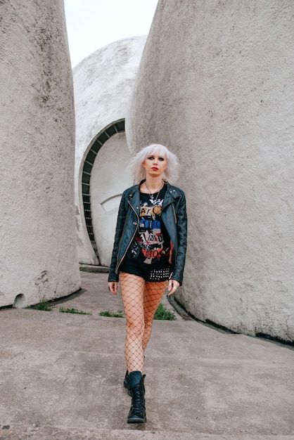 A woman in an edgy street style outfit from Shein, featuring a graphic tee, a faux leather jacket, a pleated skirt, and combat boots, accessorized with a beanie and layered necklaces.